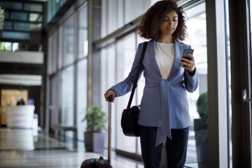 Businesswoman with curly hair, wearing a blue blazer, walking through an office hallway with a suitcase, looking at her phone.