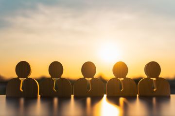 Wooden figures of businesspeople in a row with sunset in the background.