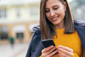 Smiling woman in a yellow sweater looking at her smartphone outdoors while wearing earbuds and a coat.