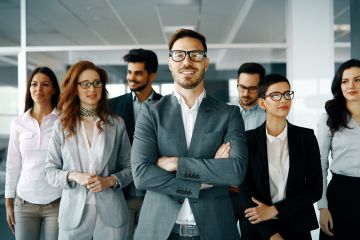 A group of smiling business professionals posing in an office, with one man in the front with crossed arms. 